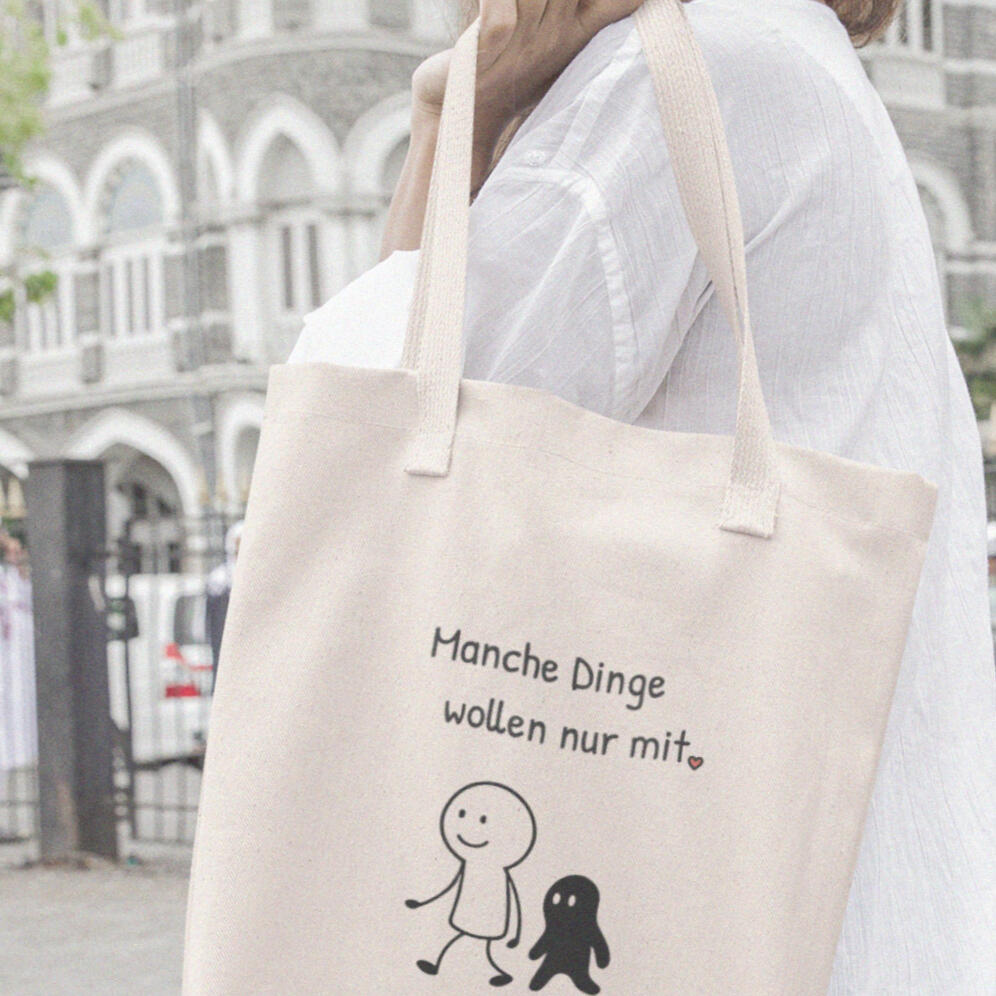 Strassenszene: Frau trägt Baumwollbeutel mit zwei minimalistischen Figuren und einem kurzen Satz "Manche Dinge wollen einfach nur mit.".