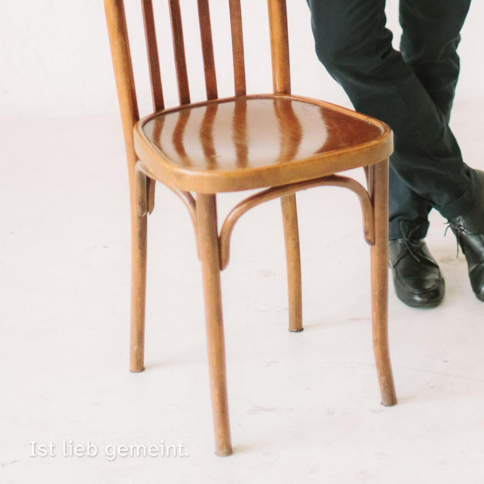 Person hält einen Holzstuhl in neutraler Studioatmosphäre, darüber der Text „Ist lieb gemeint.“ reduziert und ruhig.